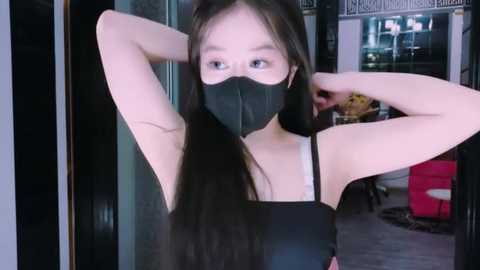 Media: Video of a young Asian woman with long black hair, wearing a black face mask and black sleeveless top, standing in a dimly lit indoor space with modern decor.