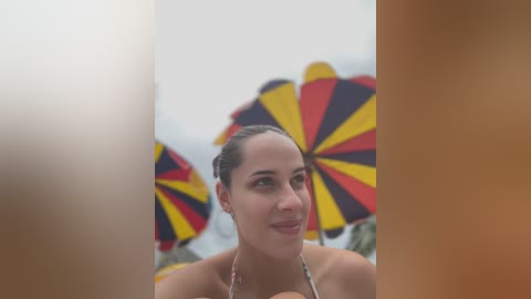 A video of a young woman with light brown skin and dark hair pulled back, wearing a bikini, holding a colorful umbrella with black, red, and yellow stripes. The background shows blurred beach chairs and an ocean view.