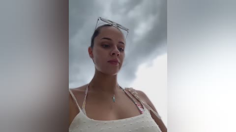 Media: Video of a young woman with dark hair, wearing sunglasses, a white floral dress, and a turquoise necklace, set against a cloudy sky background.