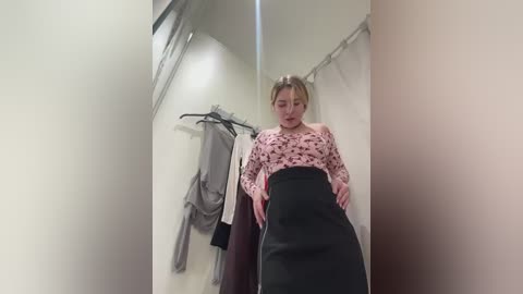 Video of a young woman with fair skin and blonde hair, wearing a pink floral blouse and black pencil skirt, standing in a fitting room with hanging clothes on the left.