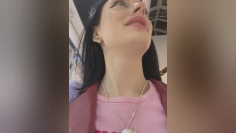 Video of a woman with long black hair, wearing a pink top and silver necklace, smiling with eyes closed, taken from a low angle.