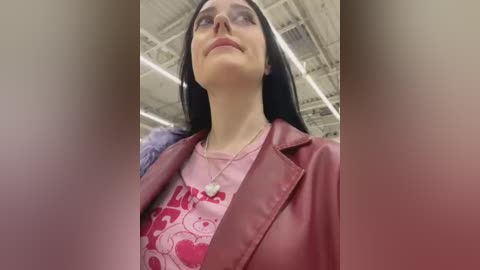 Video of a smiling woman with long black hair, wearing a pink graphic tee, red leather jacket, and glasses, standing in a warehouse with exposed pipes and lights.