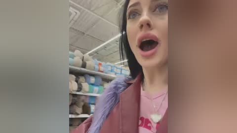 A video of a shocked woman with a purple scarf and pink shirt in a yarn store. Background shows shelves filled with colorful yarns.