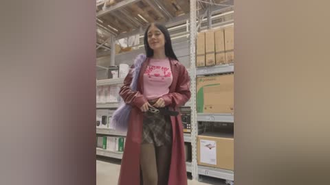 A video of a young woman with long black hair, wearing a pink graphic t-shirt and red coat, shopping in a warehouse with shelves of boxes and pallets.