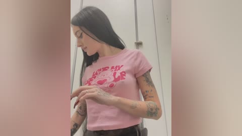 Video of a slender, tattooed woman with long black hair, wearing a pink \"Hello Kitty\" t-shirt, standing in a dimly lit, pink-walled bathroom.