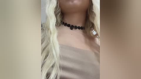 Media: Video of a woman with long, wavy blonde hair and fair skin, wearing a black floral choker and a beige off-shoulder top. The background is blurred, focusing on her upper body and neck.