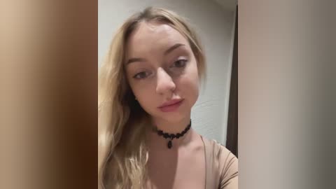 Media: Video of a young, light-skinned woman with long, wavy blonde hair and fair skin. She wears a black choker necklace and a beige top. Background includes beige walls and a wooden door frame.