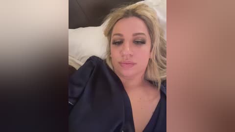 Media: Video of a blonde woman lying on a bed with white pillows, wearing a dark shirt, smiling. The background includes a blurred beige wall.