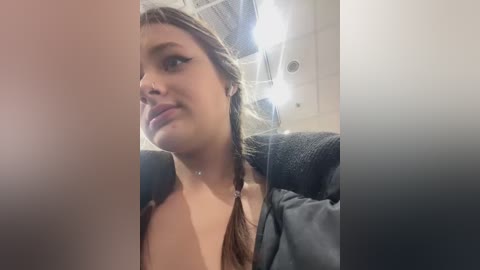 Video of a young woman with light skin and brown hair in braids, wearing a black jacket, seated on a plane, looking slightly to the right.