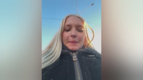 Video of a blonde woman with long hair, wearing a black jacket, looking slightly upwards with a neutral expression against a clear blue sky.