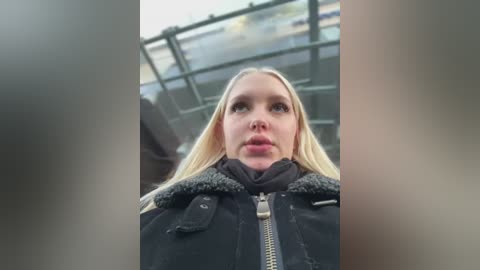 Video of a blonde woman with a serious expression, wearing a black winter coat with a fur-trimmed hood, standing on a glass bridge with a blurred cityscape in the background.