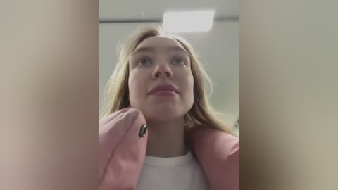 Video of a young woman with fair skin and light brown hair, wearing a white top and pink jacket, looking up with a neutral expression. The background shows a beige wall and a ceiling light.