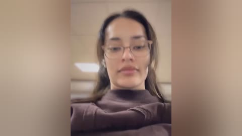 Media: Video of a young woman with fair skin, brown hair, wearing glasses, and a brown turtleneck sweater, taken from a slightly blurred, close-up angle.