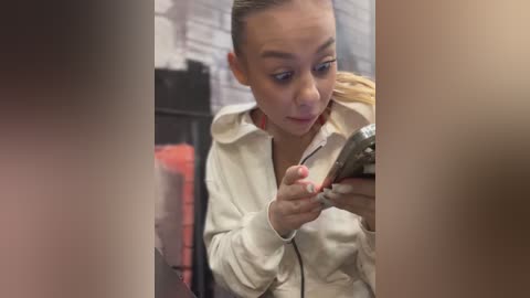 A video of a young woman with light skin, brown hair, and brown eyes, wearing a beige jacket, intently looking at her smartphone. The background features a brick wall and a red garment hanging on a rack.