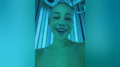 Media: A video of a smiling, topless woman with light skin and a slender physique, lying in a tanning bed with vertical slats. The room is dimly lit in a blue hue.