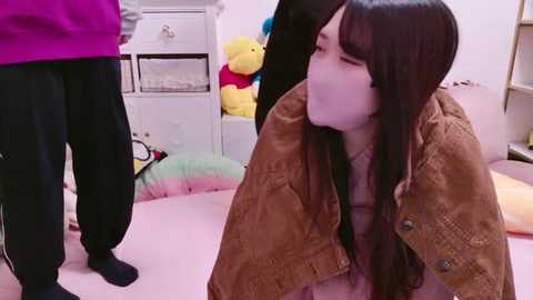 Media: Video of a young Asian woman with long black hair, wearing a brown jacket, kneeling and blowing bubbles, in a pink room with stuffed toys and a white dresser.