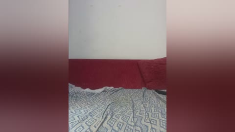 Media: A video of a bed with a red pillow and a bedspread featuring a blue and white geometric pattern. The image is framed by a gradient of red at the top and bottom, obscuring the edges.