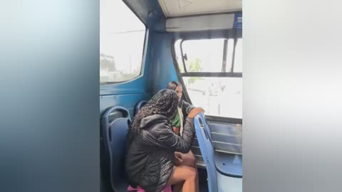 A video of a young girl with braided hair, wearing a dark jacket, seated in a blue bus, looking out the window, rain visible outside.