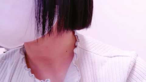 Video of a person with a short, straight black bob haircut, wearing a white, textured cardigan with scalloped edges. The background is plain white, highlighting the subject's neutral-toned attire.