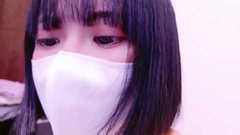 Media: Video of an Asian woman with straight, shoulder-length black hair, wearing a white face mask, showing only her eyes and mouth. The background is a blurred, neutral-toned room.