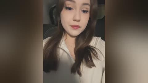 Media: Video of a young woman with fair skin and long brown hair, wearing a white blouse, sitting in a car with dark upholstery.