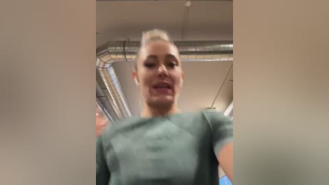 Media: A video of a blonde woman in a green shirt, taken from a low angle, showing her from mid-chest up. The background includes industrial ceiling elements and fluorescent lighting.