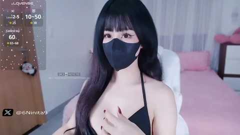 Media: Video of an East Asian woman with long black hair, wearing a black face mask and black top, sitting in a pink bed. Background includes a teddy bear and a white curtain.