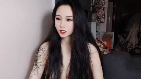 Media: Video of a fair-skinned Asian woman with long black hair, wearing red lipstick, and a black top, with tattoos on her left arm. Background shows a dimly lit room with dark furniture and a floral arrangement.