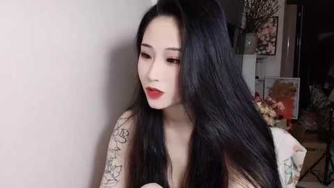 Media: Video of a young East Asian woman with long, straight black hair, fair skin, and red lipstick. She's wearing a floral-patterned top and has intricate tattoos on her arms. Background shows a dimly-lit room with floral decorations.