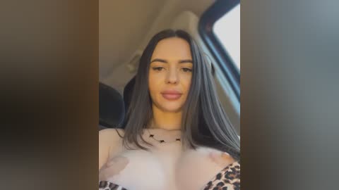 Media: Video of a young woman with long, straight black hair, light skin, and full lips, wearing a sheer leopard-print top, revealing her medium-sized breasts. She's inside a car with beige interior, blurred background.