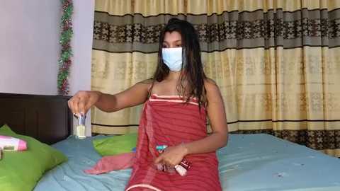 Media: Video of a South Asian woman with medium skin tone, long dark hair, wearing a red sari, blue surgical mask, and holding a syringe, standing on a bed with green and blue bedding, in a bedroom with beige curtains and a garland.