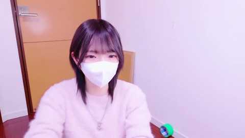 Media: Video of an Asian woman with straight black hair and bangs, wearing a white face mask and light pink sweater, indoors near a closed wooden door and a green and blue object on a wooden floor.