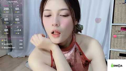 Media: Video of an Asian woman with light skin and straight dark hair, wearing a pink, sleeveless, halter-neck top, with a serene expression, in a modern, light-filled room with white curtains and wicker shelves.