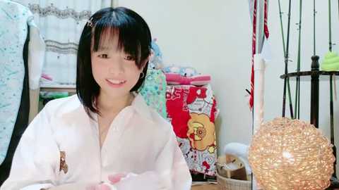 Media: Video of a young Asian woman with straight black hair, wearing a white blouse, smiling in a cluttered room with colorful clothes, a round lamp, and a bookshelf in the background.