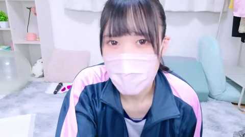 Media: Video of an Asian woman with long dark hair and a pink face mask, wearing a pink and navy jacket, sitting on a light gray carpet in a minimalist room with white walls and pastel furniture.