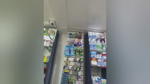 Media: Video of a pharmacy interior, showing neatly organized shelves filled with various over-the-counter medications and health products. The shelves are white with blue accents, and the lighting is bright and even.