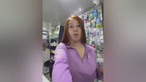 Media: Video of a middle-aged woman with red hair and pale skin, wearing a lavender zip-up jacket, working behind a pharmacy counter. Shelves behind her are packed with various medications.