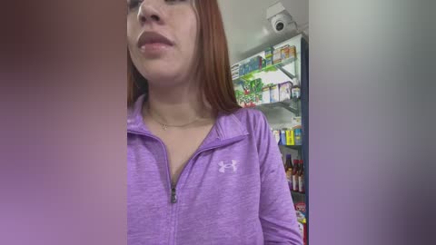 Media: A video of a young woman with fair skin, long auburn hair, and glasses, wearing a purple zip-up hoodie, standing in a well-stocked grocery store aisle.