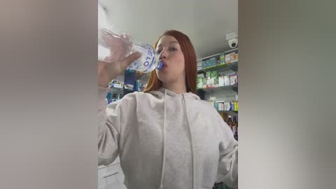 Video of a Caucasian woman with long red hair, wearing a white lab coat, drinking from a water bottle in a pharmacy aisle.