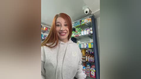 A video of a smiling, light-skinned woman with long, straight red hair, wearing a light gray hoodie, standing in a brightly lit pharmacy with shelves filled with various products and a security camera visible.