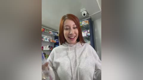 Video of a smiling, light-skinned woman with shoulder-length red hair in a white bathrobe, holding a bottle, in a cluttered pharmacy with shelves of medicines in the background.