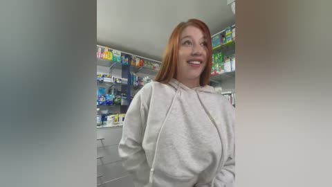 Media: Video of a fair-skinned woman with long red hair in a light grey hoodie, smiling in a pharmacy aisle filled with colorful medicine boxes.