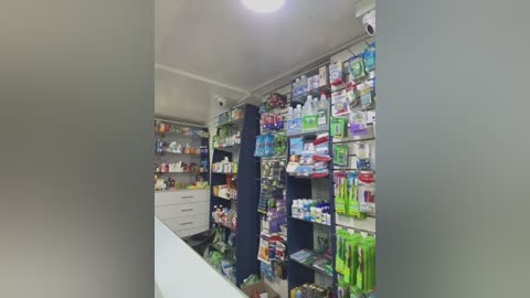 Media: Video of a brightly lit pharmacy interior with a white counter, shelves filled with colorful medication boxes, and a security camera mounted on the ceiling.