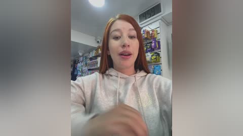 Video of a young woman with straight red hair, wearing a pink hoodie, smiling, standing in a cluttered room with shelves of colorful items.