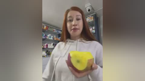 Media: Video of a young, fair-skinned woman with long red hair, wearing a white hoodie, holding a yellow lemon, standing in a brightly lit pharmacy with shelves filled with various products.