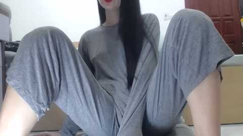 Media: Video of a slim, fair-skinned woman with long black hair, wearing loose grey pajamas, sitting with legs spread apart on a beige couch. Background features a wooden door and white walls.