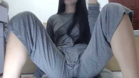 Media: Video of a young woman with long black hair, wearing grey pajamas, sitting on a beige couch, legs spread, in a domestic setting.