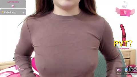 Video of a woman with long brown hair wearing a tight, brown long-sleeve top, showing her medium-sized breasts. Background includes a bed, a pink and white pillow, and a digital clock.