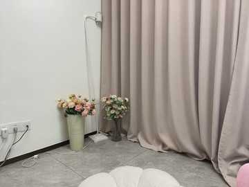 Video of a minimalist room with beige curtains, a white wall, and a gray floor. Two flower vases with mixed pink and white flowers stand on either side of a white lamp.