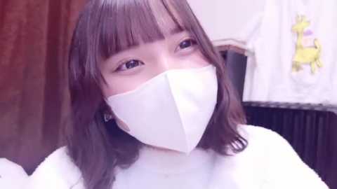 Media: Video of a young East Asian woman with shoulder-length brown hair, wearing a white face mask, white sweater, and a floral-patterned white apron. Background includes a wooden door and a dark countertop.